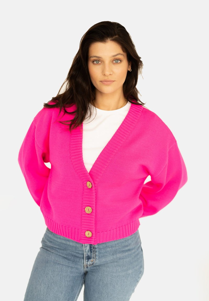 Fuksjowy, oversize kardigan z prążkowanym dekoltem i mankietami, z drewnianymi guzikami z przodu; zestawiony z jasnoniebieskimi dżinsami.