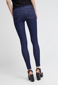 Jeans de mezclilla oscuros y ajustados, con dos bolsillos traseros que presentan un patrón de costura distintivo y un detalle con cremallera en la cintura.