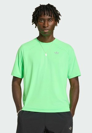 Young man with dark skin and dreadlocks wearing a bright green Adidas t-shirt and black shorts, standing with hands in pockets.