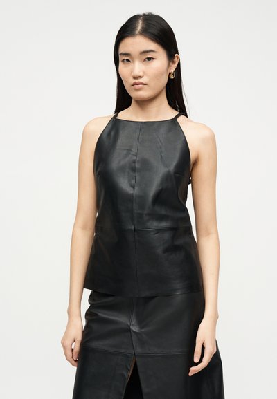 Γυναίκα με μακριά μαύρα μαλλιά που φοράει αμάνικο μαύρο δερμάτινο τοπ και ταιριαστή φούστα με σχίσιμο μπροστά, στέκεται μπροστά σε απλό λευκό φόντο.