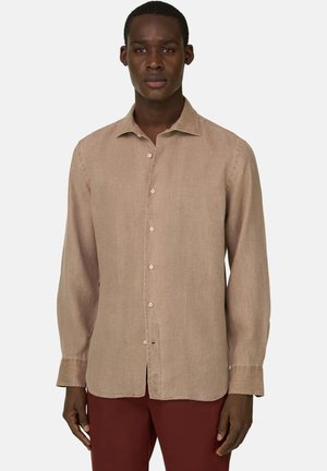 Mann trägt ein beiges langärmeliges Hemd zum Knöpfen und weinrote Hosen, steht vor einem einfarbigen hellen Hintergrund.