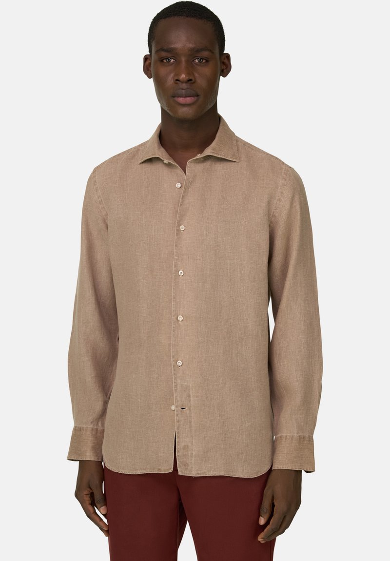 Homme portant une chemise beige à manches longues et un pantalon bordeaux, debout devant un fond clair uni.