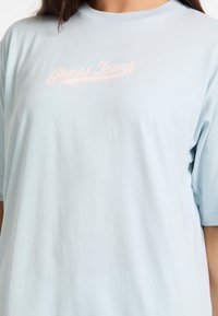 Lyseblå bomulds t-shirt med rund hals og korte ærmer, med "Guess Jeans" logo i pink skrift over brystet.