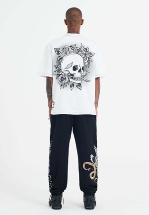 Camiseta blanca de gran tamaño con un gráfico negro de calavera y rosa en la parte posterior. Combinada con pantalones de chándal negros que presentan coloridos diseños de serpiente y alas.