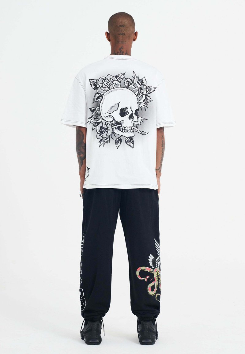 Camiseta blanca de gran tamaño con un gráfico negro de calavera y rosa en la parte posterior. Combinada con pantalones de chándal negros que presentan coloridos diseños de serpiente y alas.