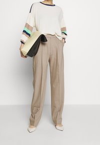 Krämfärgad tröja med flerfärgade randiga ärmar, ihop med beige plisserade byxor. Modellen håller en tvåtonad väska. Vita spetsiga skor.