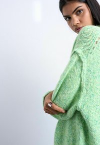 Jeune femme portant un pull en maille texturée vert clair, ajustant la manche avec une main ornée de deux bagues argentées, fond blanc.