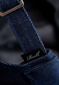 Correa de denim con herrajes plateados y etiqueta negra que presenta texto blanco. Detalle en primer plano que muestra la textura y las costuras del tejido.