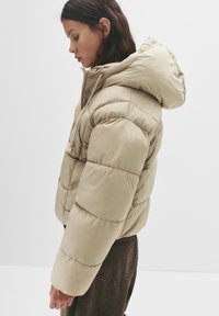 Giacca puffer beige con cappuccio, caratterizzata da un design trapuntato e texture a coste. Modello mostrato di lato, che indossa pantaloni scuri.