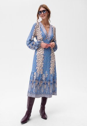 Langes Kleid mit tiefem V-Ausschnitt, blau-beigem Blumenmuster, puffärmeln und gemustertem Saum. Kombiniert mit hohen bordeauxfarbenen Stiefeln.