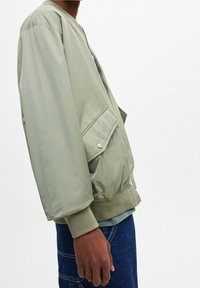 Bomber jacket verde chiaro con zip frontale, polsini e orlo a coste, e tasca laterale; realizzato in materiale liscio e leggermente lucido.