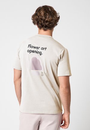 Uomo che indossa una maglietta beige con la scritta "Apertura arte floreale." e un disegno astratto di un fiore stampato sul retro, su sfondo uniforme.