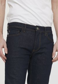 Donkerblauwe spijkerbroek met oranje stiksels, vijf zakken en een metalen knoopsluiting. Het materiaal lijkt duurzaam met een klassieke denim textuur.
