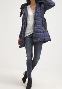 Chaqueta de abrigo azul marino con capucha de pelo sintético, cierre de cremallera y botón, bolsillos laterales y un acento de cinturón, combinada con jeans ajustados y botas de tobillo.