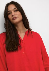 Röd linneblus med mandarin krage, med en knappslå och långa ärmar, uppvisad på en modell mot en neutral bakgrund.