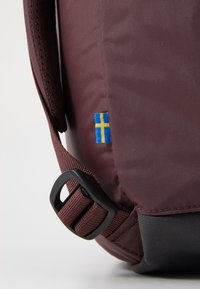 Esquina de bolsa de tela color marrón con un pequeño parche de la bandera sueca. Correa de hebilla negra sujeta al costado, con textura suave.