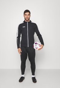 Zwarte sportjack met witte accenten, bijpassende broek en zwarte voetbalschoenen. Persoon houdt een witte voetbal vast met kleurrijke details.