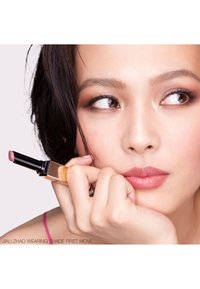 Jeune femme aux cheveux foncés tenant un rouge à lèvres mauve près de son visage, regardant sur le côté avec un maquillage naturel et des lèvres rose pâle.