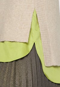 Gestapelte Kleidungsdetails mit einem beigen, gerippten Pullover, einem limonengrünen Shirt mit abgerundetem Saum und einem gefältelten, dunkelbraunen Rock.