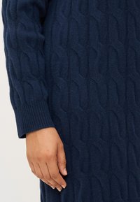 Eine Nahaufnahme einer Hand, die auf einem strukturierten, dunkelblauen gestrickten Kleidungsstück ruht, das ein Zopfmuster und gerippte Bündchen zeigt.