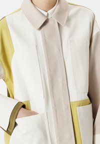 Veste bicolore en crème, beige et vert olive. Comprend un col pointu, des poches frontales et des panneaux texturés contrastants.