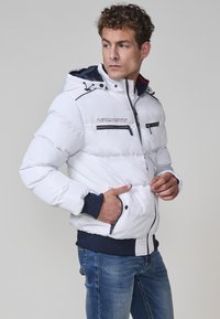 Chaqueta acolchada blanca con capucha azul marino, que cuenta con cremalleras y un logo en el pecho. Puños y dobladillo acanalados con una textura suave y brillante.