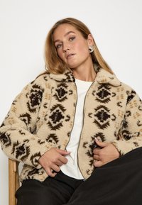 Beige vilnas jakas ar brūniem ģeometriskiem rakstiem, ar rāvējslēdzēju un mīkstu tekstūru. Valkāta virs baltās blūzes, saskaņota ar melnām biksēm.