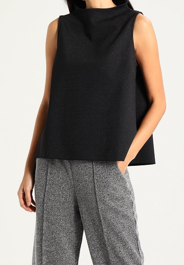 Femme portant un haut noir sans manches à col montant et un pantalon gris à jambes larges, une main dans la poche, sur un fond uni.