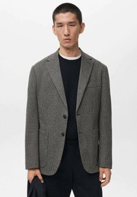 Grau kariertes Blazer mit strukturierter Passform, einreihigem Design, Eingrifftaschen und zwei Vorderfächern. Über einem schwarzen Pullover getragen.