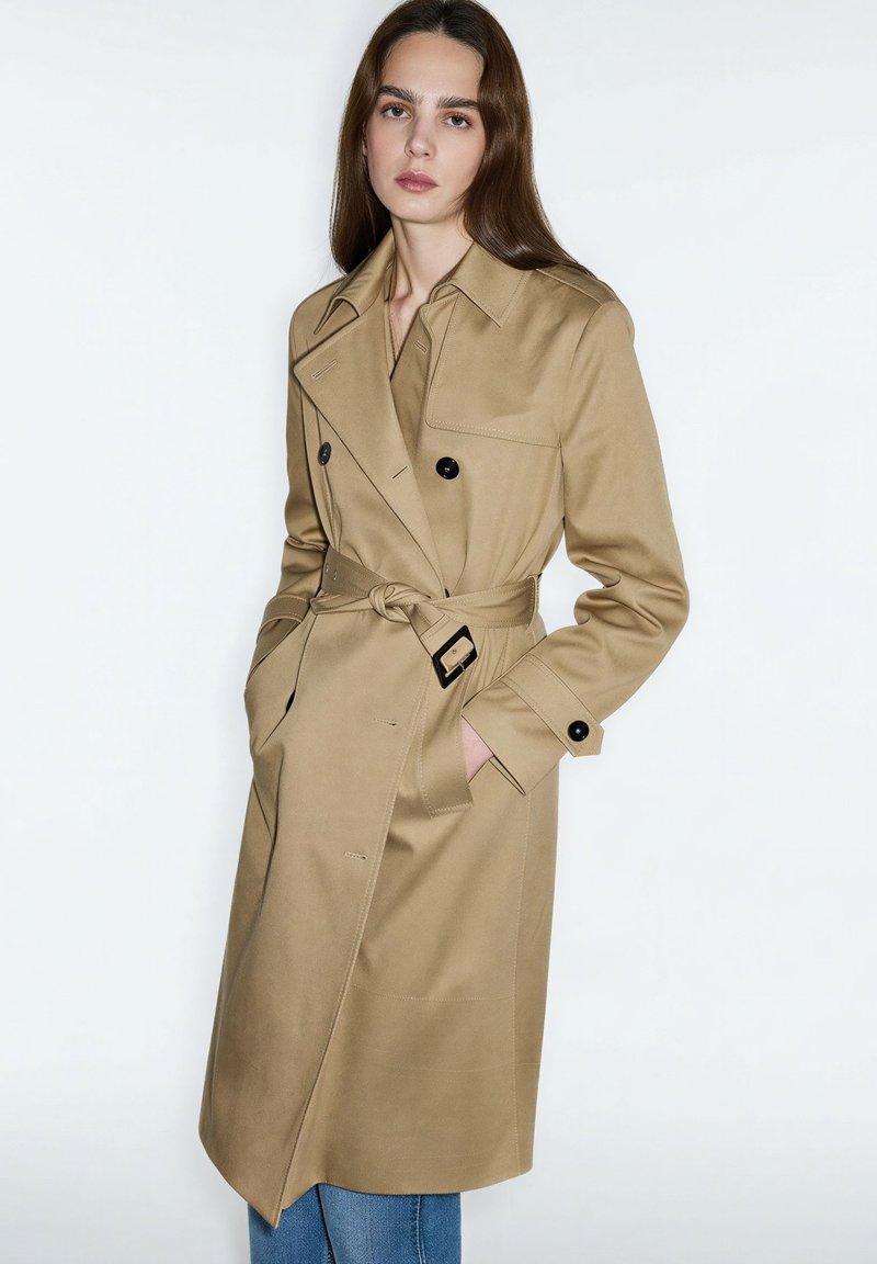 Woman wearing a beige belted trench coat with black buttons and blue jeans, standing with hands in coat pockets against a white background.