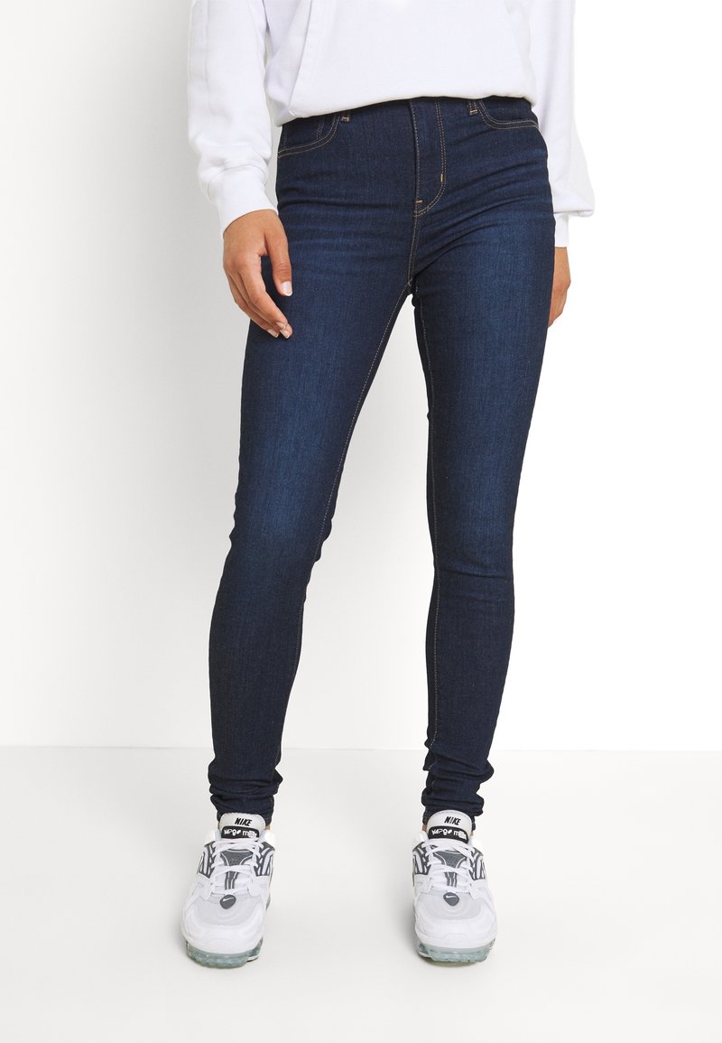 Personne portant un jean skinny bleu foncé, des baskets blanches Nike avec des détails noirs, et un haut blanc à manches longues, debout devant un fond blanc.
