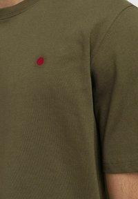 Olive zelené bavlněné tričko s kruhovým výstřihem a krátkými rukávy. Na levé části hrudi se nachází malé červené vyšívané kolečko.