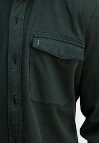 Dunkelgrüne Knopfbluse mit einer Brusttasche, ausgestattet mit einem kleinen silbernen Logo und einem weichen, strukturierten Stoff.