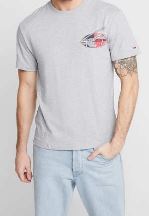 Mann trägt hellgraues T-Shirt mit kleinem Brustmotiv und hellblaue Jeans, rechte Hand in der Tasche, sichtbares Tattoo am linken Arm.