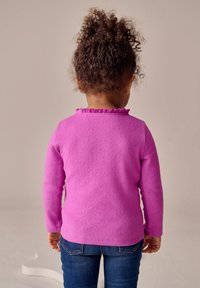 Suéter de manga larga color fucsia con un acabado texturizado y cuello con volantes, usado con jeans de mezclilla azul, visto desde atrás.