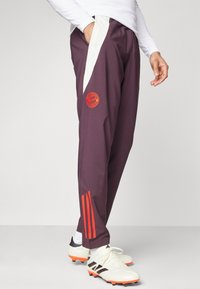 Paarse sportbroek met witte zijpanelen, FC Bayern München-logo in het rood en drie rode strepen op het onderbeen. Draagt witte voetbalschoenen.