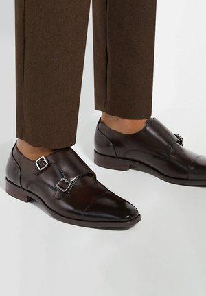 Ciemnobrązowe skórzane eleganckie buty z podwójnym paskiem typu monk, noszone z brązowymi spodniami garniturowymi na białym tle.