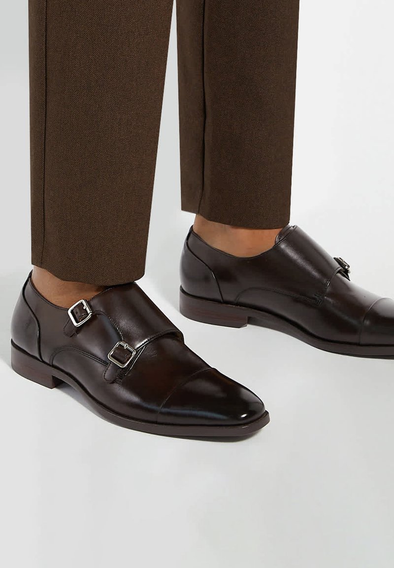Chaussures habillées en cuir marron foncé à double boucle monk, portées avec un pantalon de costume marron sur fond blanc.