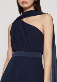 Robe bleu marine avec un design à une épaule, présentant un tissu drapé, une taille ajustée et une jupe fluide avec un overlay transparent sur l'épaule.