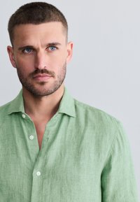 Chemise en lin vert clair avec un col, présentant une finition texturée et des boutons blancs. La chemise a une coupe décontractée et une ouverture subtile au niveau du cou.