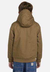 Brun hættejakke med en glat tekstur, elastiske manchetter og en tætsiddende kant. Bæres over blå denim jeans med et bagpatch-logo.