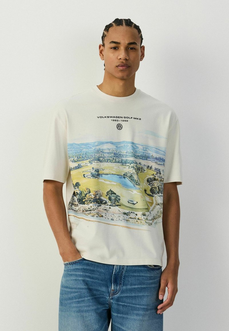 Giovane uomo con capelli intrecciati che indossa una maglietta bianca con grafica Volkswagen Golf MK2 e jeans blu, in piedi davanti a uno sfondo bianco uniforme.