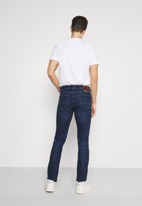 Mörkblå denimjeans med slim fit, med bakfickor och en brun läderlapp på midjan, kombinerat med en enkel vit T-shirt.