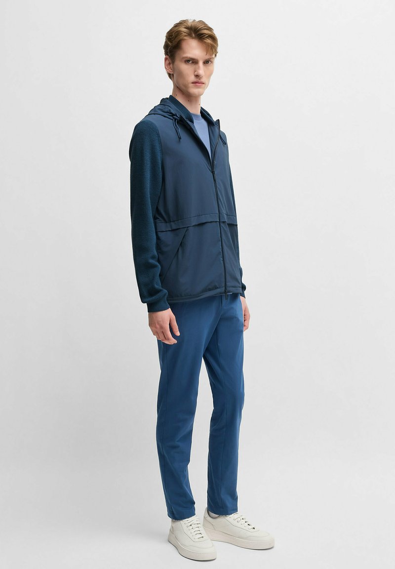 Marineblauer Jacket mit Reißverschluss, Kapuze und strukturierten Ärmeln, kombiniert mit einfarbigen blauen Hosen und weißen Turnschuhen.