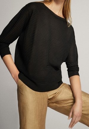 Femme portant un pull à manches longues noir côtelé rentré dans un pantalon marron clair texturé, posant avec une main dans la poche.