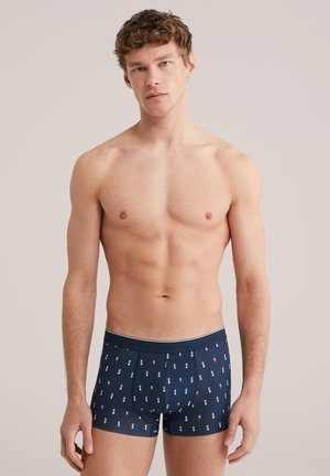 Mladi moški s kodrastimi lasmi, oblečen v temno modre bokserice z vzorcem majhnih belih in rdečih simbolov kart, stoji pred enobarvnim ozadjem.