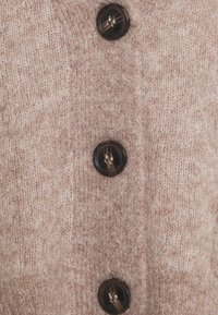 Beige pletený cardigan s čtyřmi tmavě hnědými knoflíky, texturovaná tkanina s jemným, chlupatým vzhledem a přírodními švy podél okrajů.