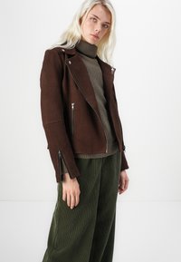 Brown suede motorcycle jacket with asymmetric zipper, silver hardware, and zippered cuffs, worn over a textured olive turtleneck and green pants.