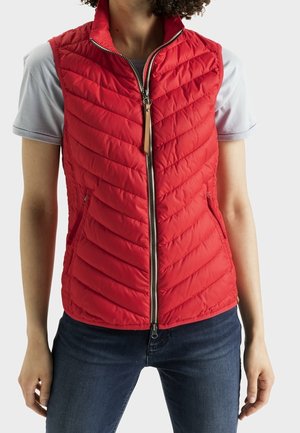 Personne portant une veste matelassée rouge sans manches zippée sur une chemise gris clair à manches courtes et un jean bleu foncé sur un fond uni.