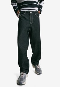 Person iført store sorte jeans med hvid syning, grå og hvide sneakers samt en sort og hvid stribet langærmet skjorte, står med hænderne i lommerne.